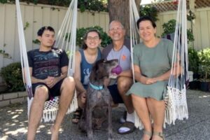 Buckshot with purple toy in his mouth, under a tree, sitting in front of 4 people in his new family, two of whom are sitting in rope swing seats