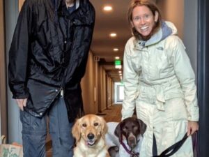 Remington next to his golden retriever sibling, with new dad and mom in raincoats in a hallway